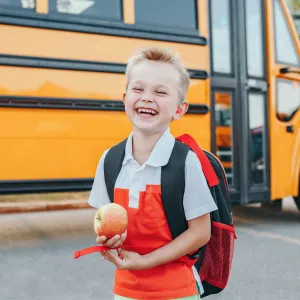 A Small Boy Laughs as He Gets Off a School Bus with an Apple in Hand.