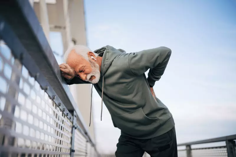 A senior man hunching over against a fence with his hand resting on his back in pain.